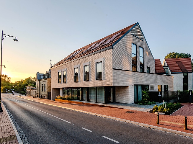 Šiuolaikinis seimos klinika pastatas miesto gatvėje – modernios architektūros, su šviesių plytų fasadu, dideliais langais ir tvarkinga aplinka, atspindinti patikimą ir šiuolaikišką sveikatos priežiūros įstaigą.